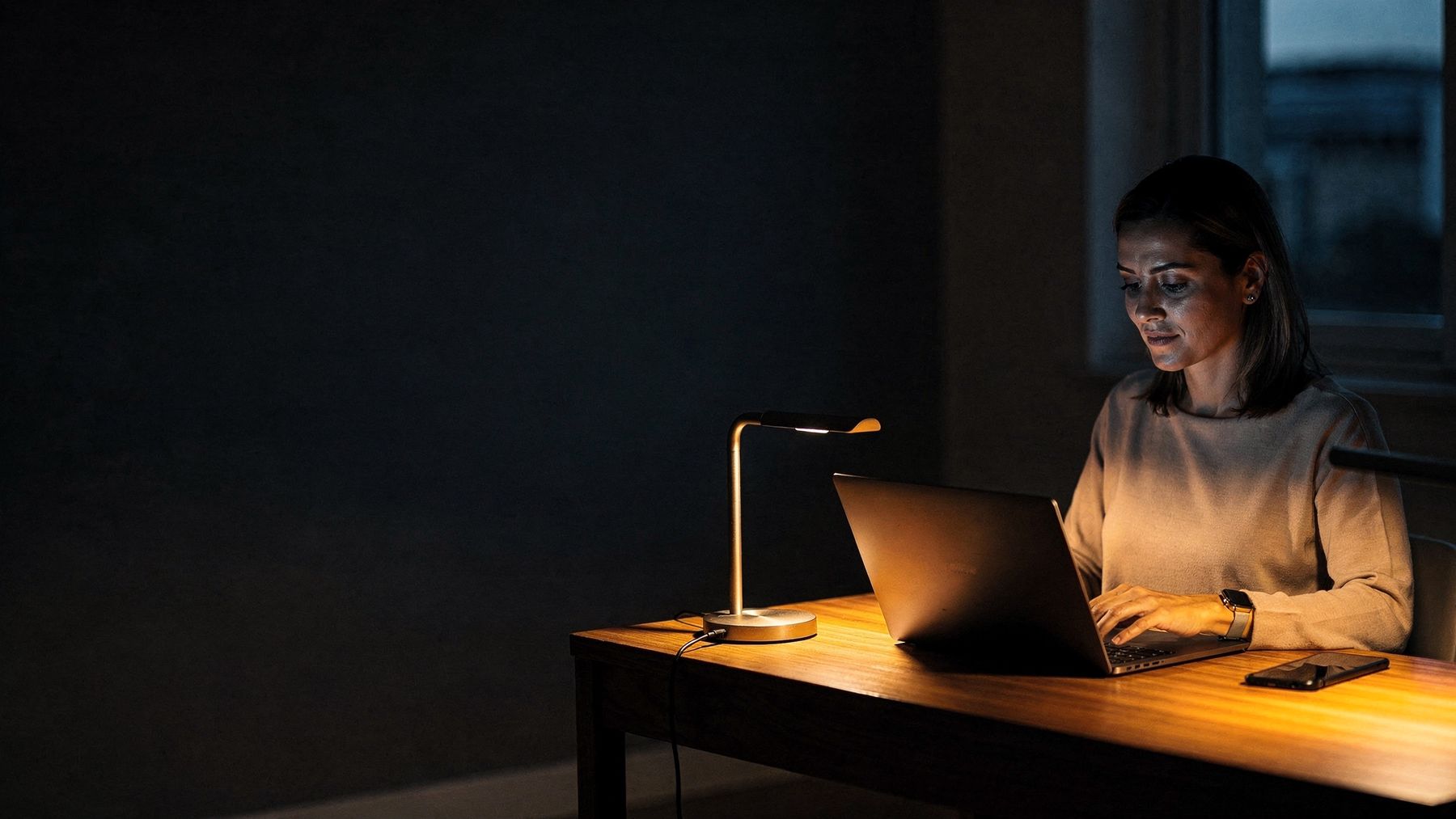 Mulher trabalhando à noite em uma mesa de madeira, com notebook, celular e uma luminária acesa